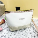 Bolso organizador de maquillaje blanco con base texturizada beige, cremallera dorada y correa rosa, ideal para viajes.