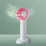 Ventilador de mano blanco con aro rosa, sin aspas, expulsa aire con hojas verdes de fondo.