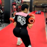 Mujer con cinturón Hipsthrust rosa para glúteos, en gimnasio, realizando ejercicio con pesas.