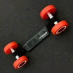 Pesas de mano con extremos rojos, conectadas por un cinturón negro de STTRIKER sobre superficie oscura con motas.