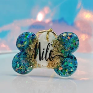 Placa de mascota en forma de hueso azul con glitter dorado y nombre "Milo" en negro.