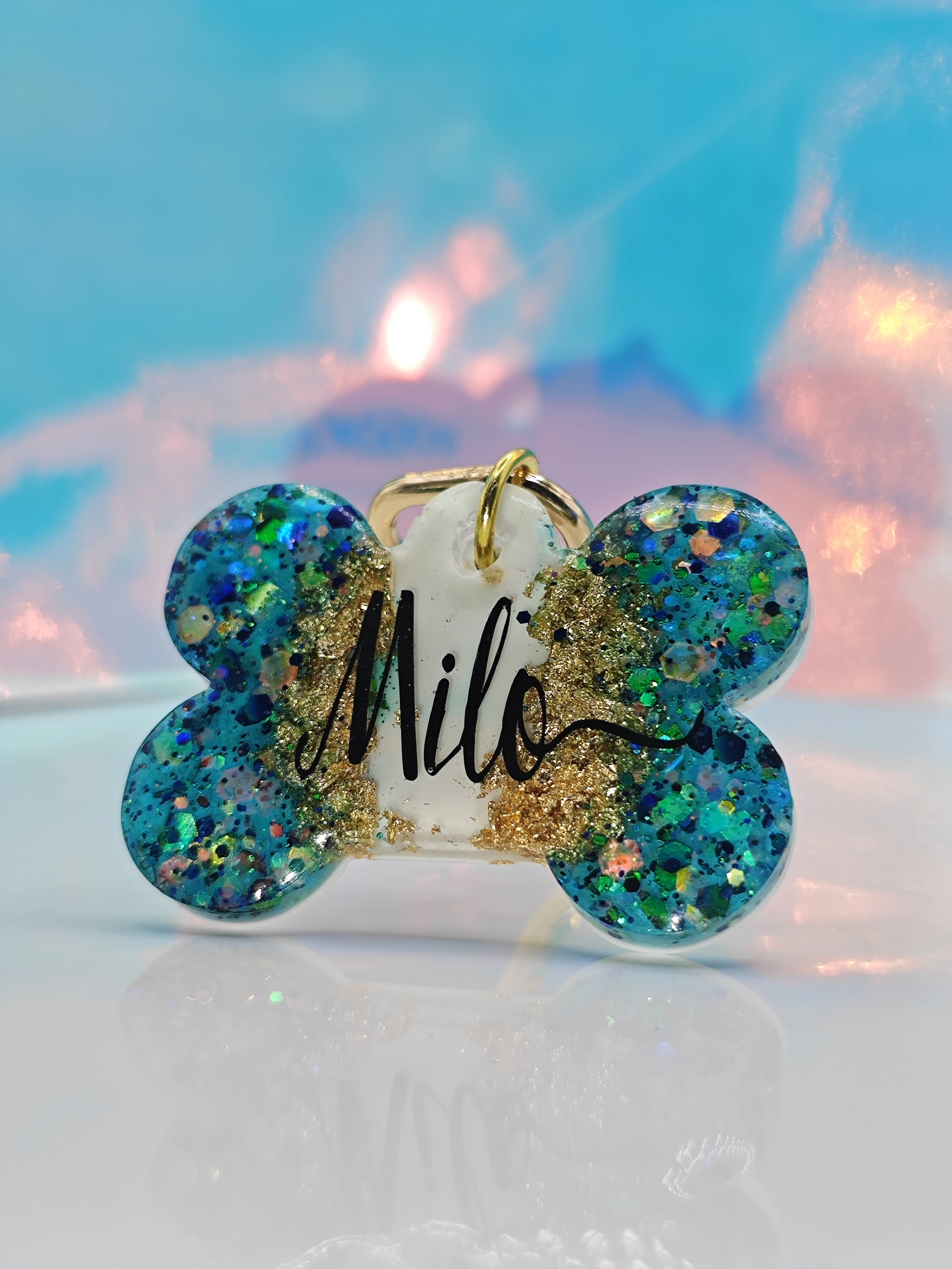 Placa de mascota en forma de hueso azul con glitter dorado y nombre "Milo" en negro.
