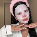 Mujer con mascarilla facial rosa y diadema, usando pijama, detalles de joyas y uñas largas.