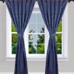 Cortinas Jacquard azul marino con patrón circular y ojales naranjas, decoran una ventana con vista a árboles.