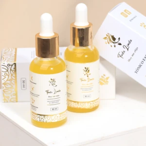 Dos frascos de vidrio de 30 ml con suero amarillento, tapas doradas con gotero y etiquetas blancas con detalles dorados, junto a cajas del mismo diseño.