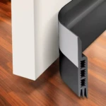 Guardapolvo gris flexible en forma de U, se adhiere a la parte inferior de una puerta blanca sobre piso de madera.