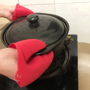 Manos con guantes de silicona rojos tipo pinza cangrejo sujetan una olla negra con tapa sobre una estufa de vidrio, protegiendo del calor al cocinar.