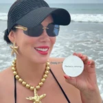 Mujer sonríe en la playa sosteniendo producto Marbella Beauty Matificante Facial, luce sombrero, gafas, collar y aretes dorados de estrella.