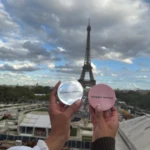 Dos compactos de matificante facial Marbella Beauty, uno transparente con reflejo del cielo parisino y otro rosa, Eiffel de fondo.