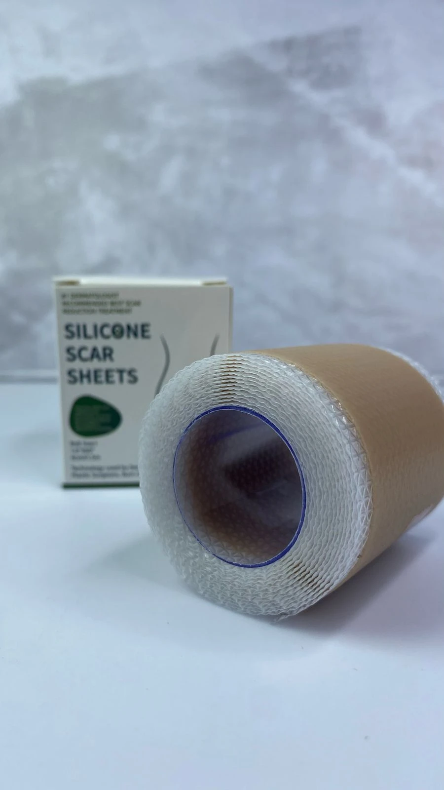 Cinta de silicona para cicatrizar en rollo color piel junto a su caja, con textura visible y borde azul en el centro del eje del empaque.