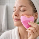 Limpiador facial en forma de corazón rosa con bordes dorados y cerdas suaves, usado por mujer en rutina de cuidado.