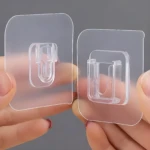 Dos ganchos adhesivos transparentes, uno a la izquierda y otro a la derecha, sostenidos por dedos.