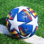 Balón de fútbol profesional #5 azul con estrellas blancas, diseño multicolor, sobre césped artificial y línea de banda blanca.