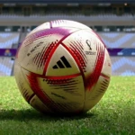 Balón de fútbol profesional Adidas Al Rihla #5 sin costuras, blanco con detalles rojos, sobre césped de estadio.