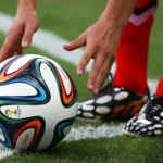 Balón de fútbol profesional Adidas Brazuca #5 con diseño colorido, sobre césped y al lado de la línea blanca, listo para ser pateado por futbolista.