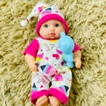 Muñeco bebé con pijama de cupcakes y osito de plástico azul sobre alfombra peluda.