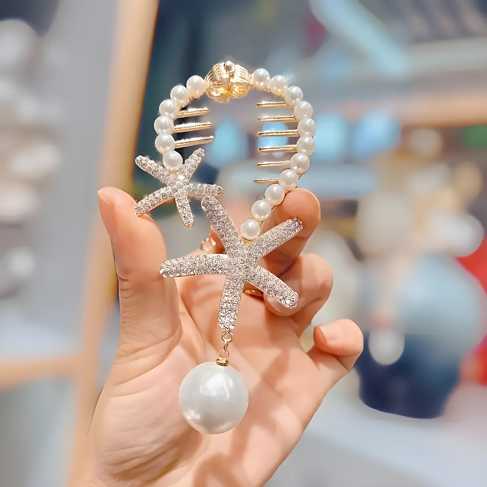 Pinza para cabello con estructura circular de perlas blancas, adornada con dos estrellas de mar brillantes en pedrería y un dije de perla grande colgante.