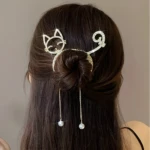 Peinado de moño con pinza metálica dorada de diseño felino, decorada con una silueta de gato y cola rizada, que incluye dos delicados dijes con perlas.