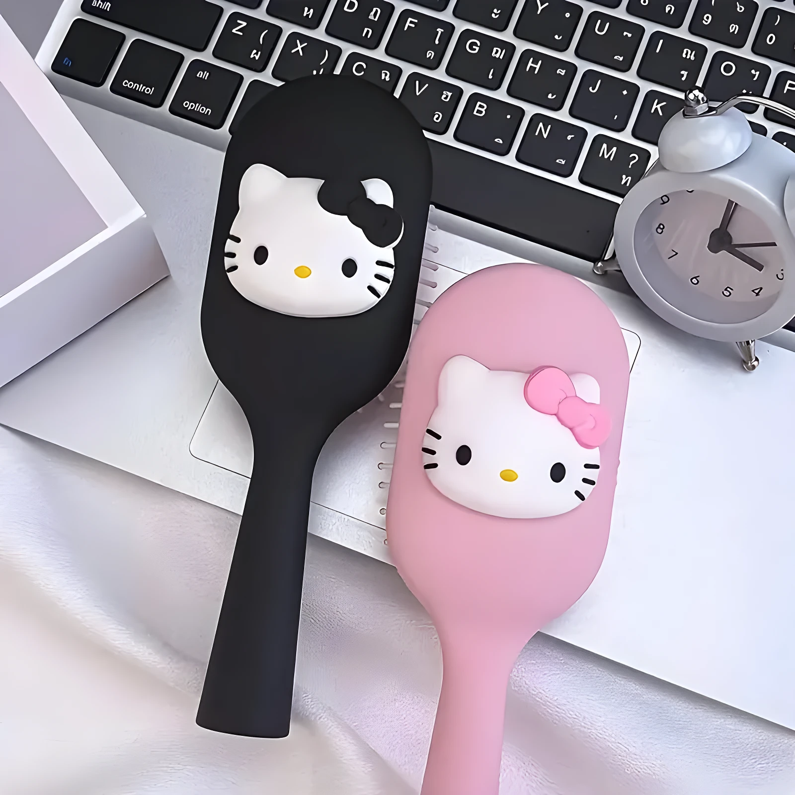 Set de dos cepillos para cabello tipo paleta con figuras de Hello Kitty, uno negro y uno rosa, sobre un teclado de computador junto a un reloj de mesa.