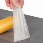 Cinta doble faz transparente con malla de refuerzo siendo retirada de un rollo amarillo, ideal para pegar objetos sin dañar superficies.