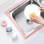 Cinta selladora adhesiva impermeable rosa y gris para borde de lavaplatos, con plato blanco y esponja amarilla.
