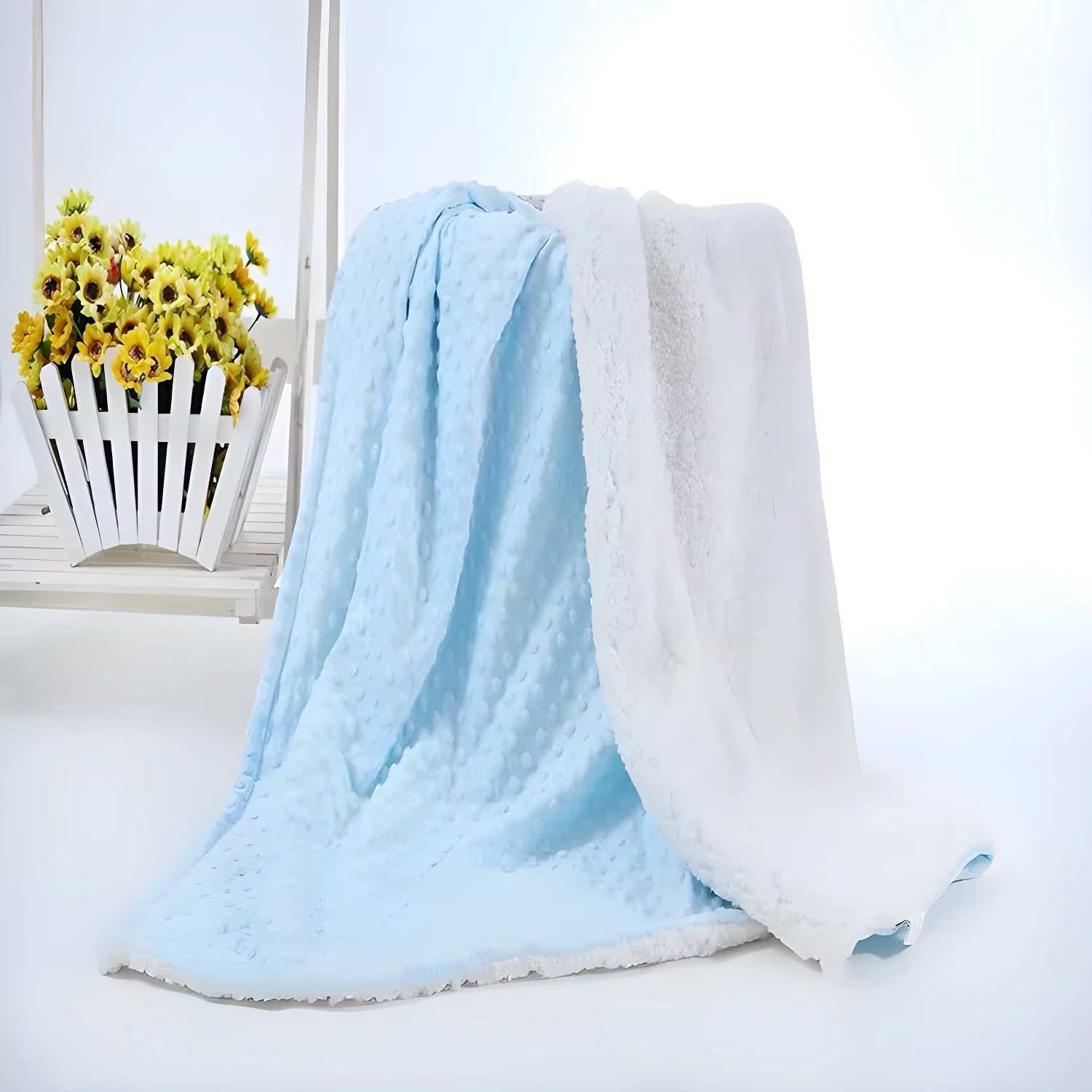Cobija térmica azul con textura de puntos, volteada a una cara blanca afelpada. Cerca, un cesto con flores amarillas.