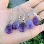 Cinco colgantes de amatista natural morada en forma de lágrima con cadenas de plata, sostenidos en una mano.