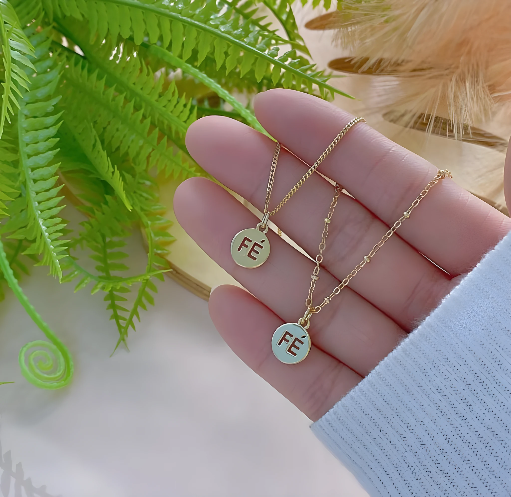 Set de 2 collares dorados con colgante redondo y la palabra "FE", uno más arriba que el otro, sobre mano con plantas de fondo.