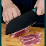 Cuchillo negro de chef cortando carne cruda en tabla de madera.
