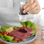Dispensador de aceite con brocha aplicando salsa a bistec crudo servido con lechuga y tomate.