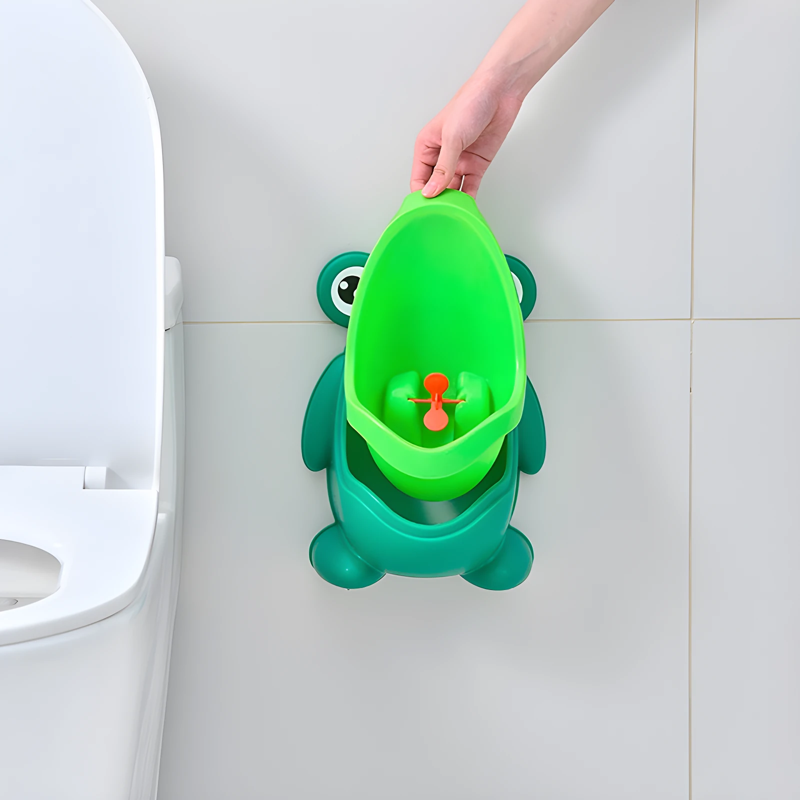 Orinal sapito verde para niños, diseñado para pared, con huevito naranja. Al lado, parte de sanitario blanco.