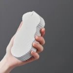 Esponja exfoliante 3D blanca con textura de escamas, sostenida en mano.