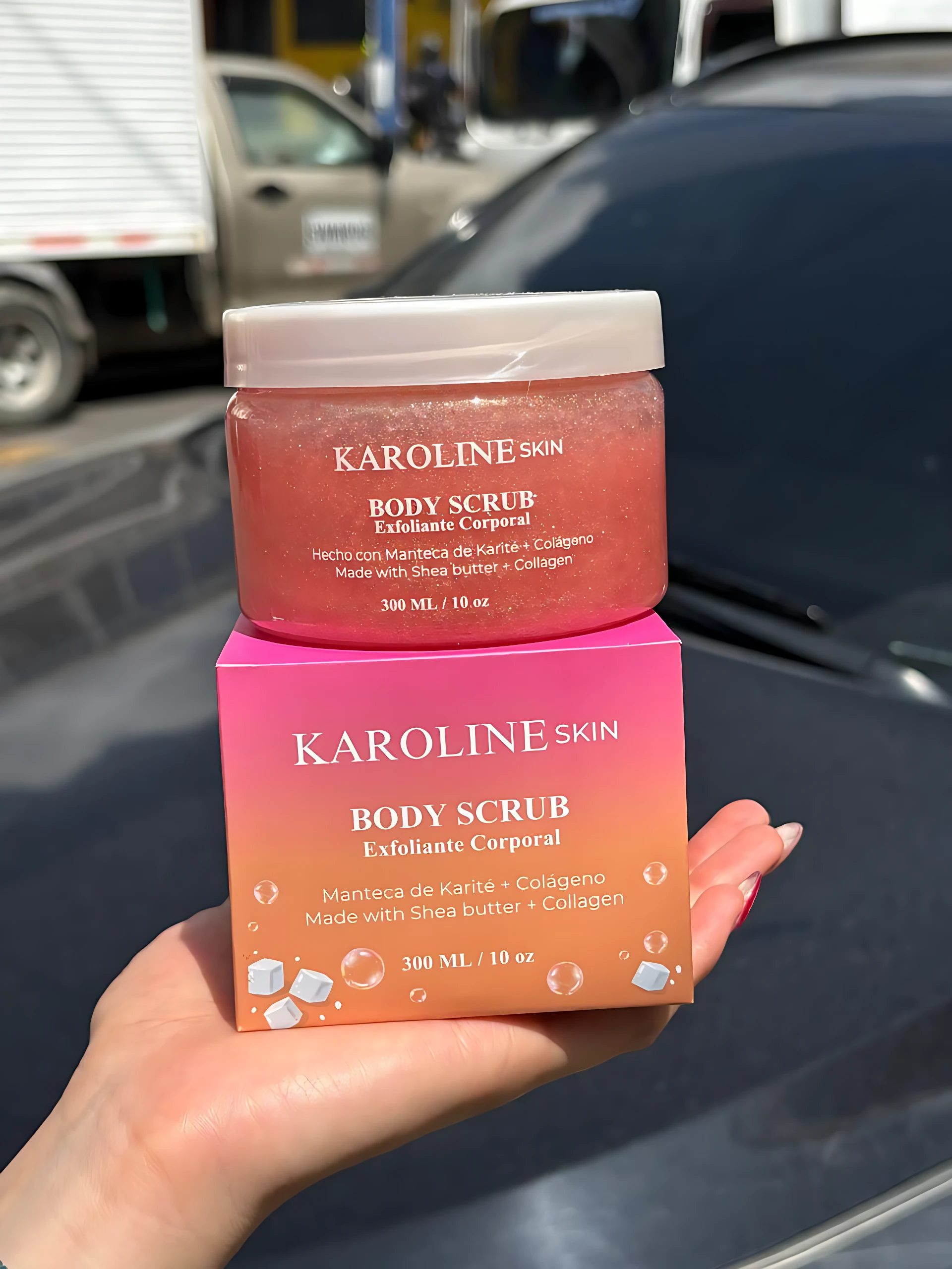 Exfoliante corporal Karoline Skin 300 ML, fragancia a rosas, en envase rosa y su caja, sostenido por una mano.