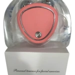 Gimnasia Facial Estimulante Electro en forma de corazón color coral con botón de encendido metálico, sobre caja blanca con texto.