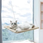 Gato blanco de ojos azules en hamaca beige para ventana con ventosas, observando el exterior.
