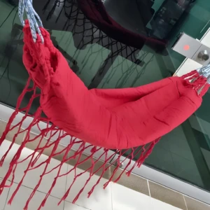 Hamaca de bebé roja con flecos y cuerdas azules, colgada junto a una puerta de vidrio.