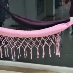 Hamaca de bebé rosa con flecos de crochet y cuerdas de anclaje grises, colgada sobre vidrio oscuro con reflejos.