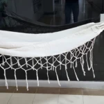 Hamaca de bebé blanca con flecos, sostenida por manos, colgada en interior moderno.