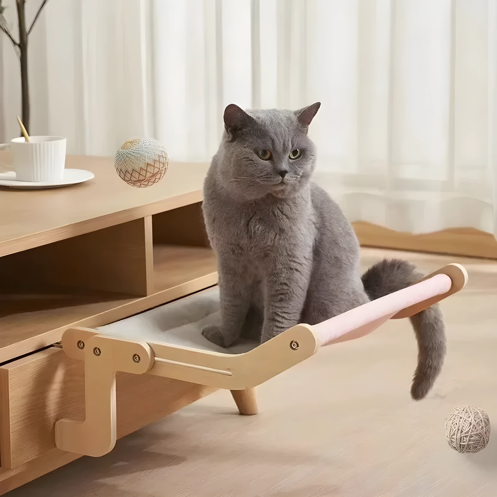 Gato gris sentado en hamaca rosa de gato para sofá, incluye pelota de cuerda, ideal para descanso seguro.