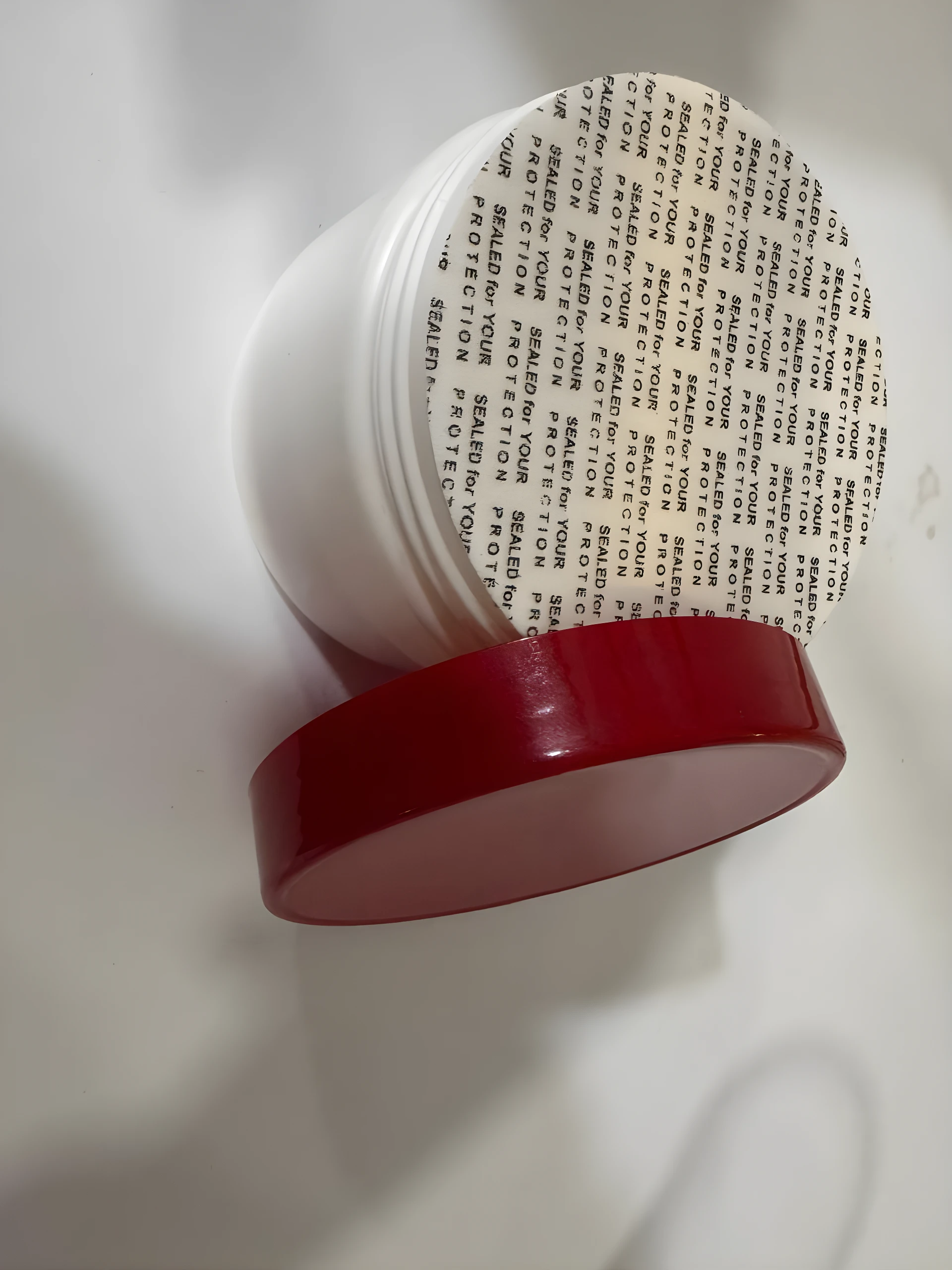 Envase blanco de inositol en polvo con tapa roja retirada, mostrando un sello de seguridad protector sobre la boca del frasco sobre fondo blanco.