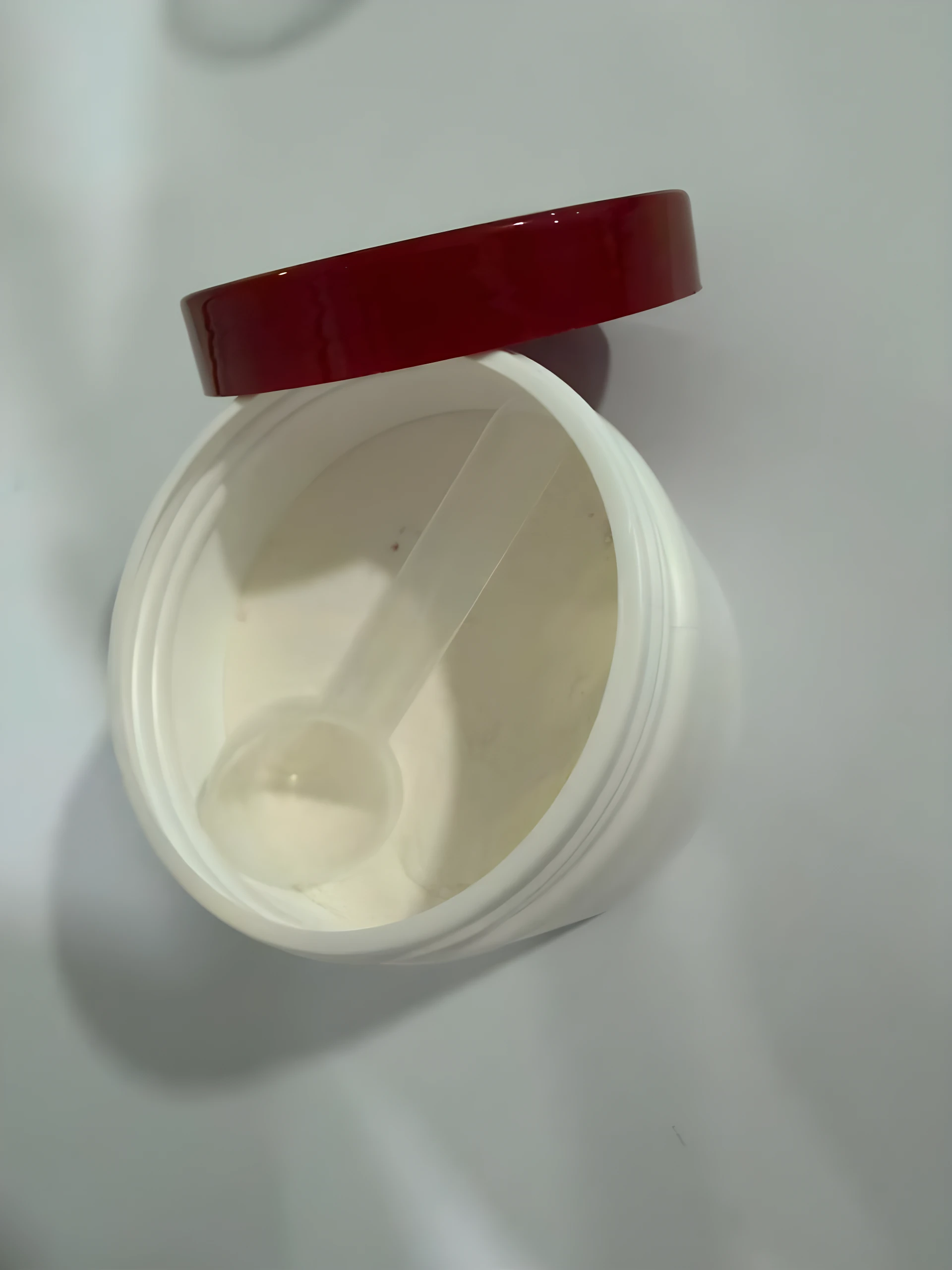 Tarro de inositol en polvo con tapa roja abierta, incluye una cuchara medidora de plástico transparente en su interior sobre el suplemento blanco.