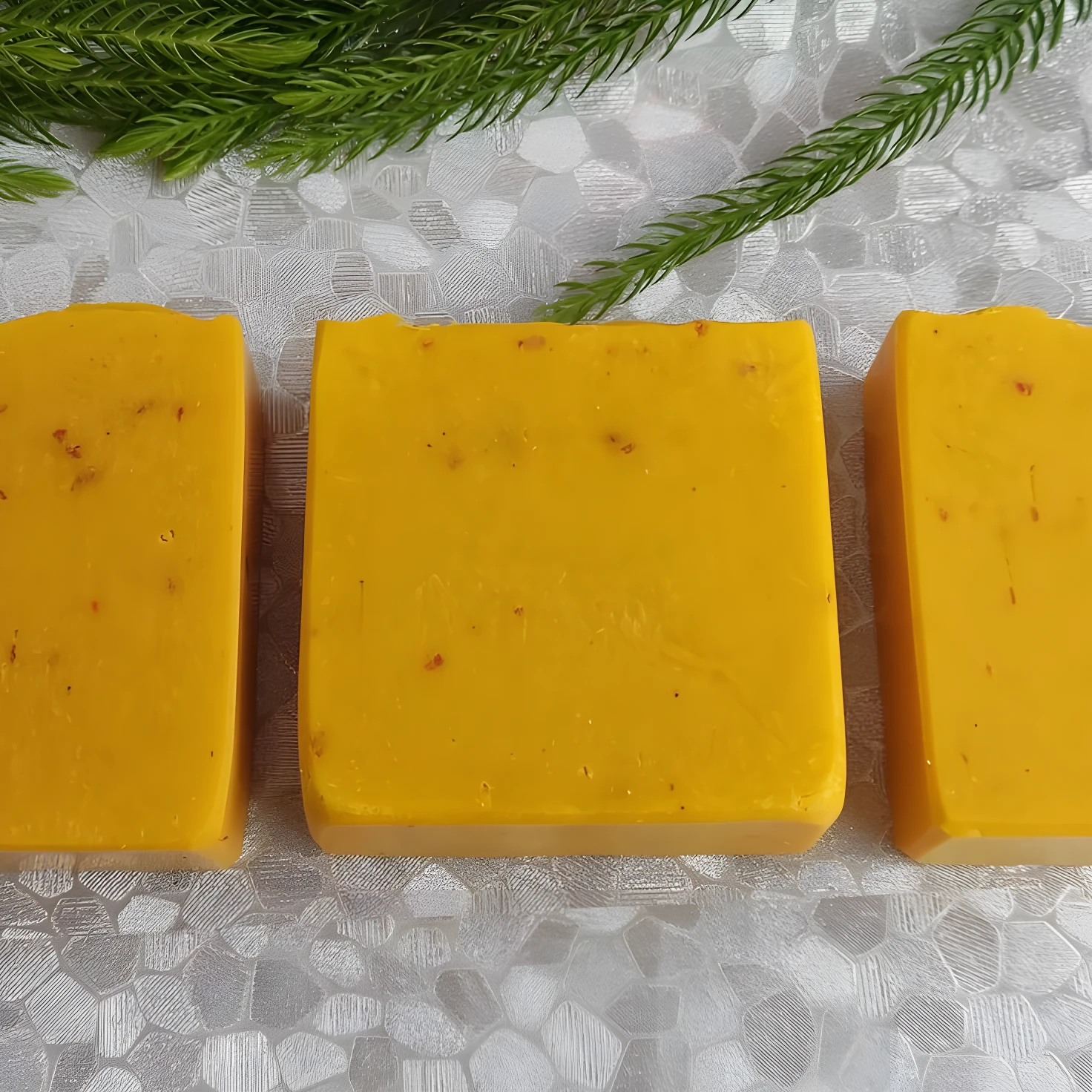 Tres barras rectangulares de jabón de cúrcuma amarillo brillante con vetas rojizas sobre fondo blanco texturizado y ramas verdes.