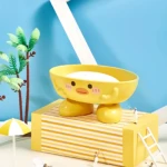 Jabonera amarilla infantil con diseño de pato, patas, detalles faciales tiernos, sobre caja a rayas.