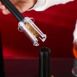 Sacacorchos de aire a presión sacando corcho de botella de vino, con cortador de cápsula y copa a un lado.