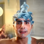 Hombre con espuma en la cabeza y un muñeco de jabón; tiene tatuajes de mariposa y ramas.