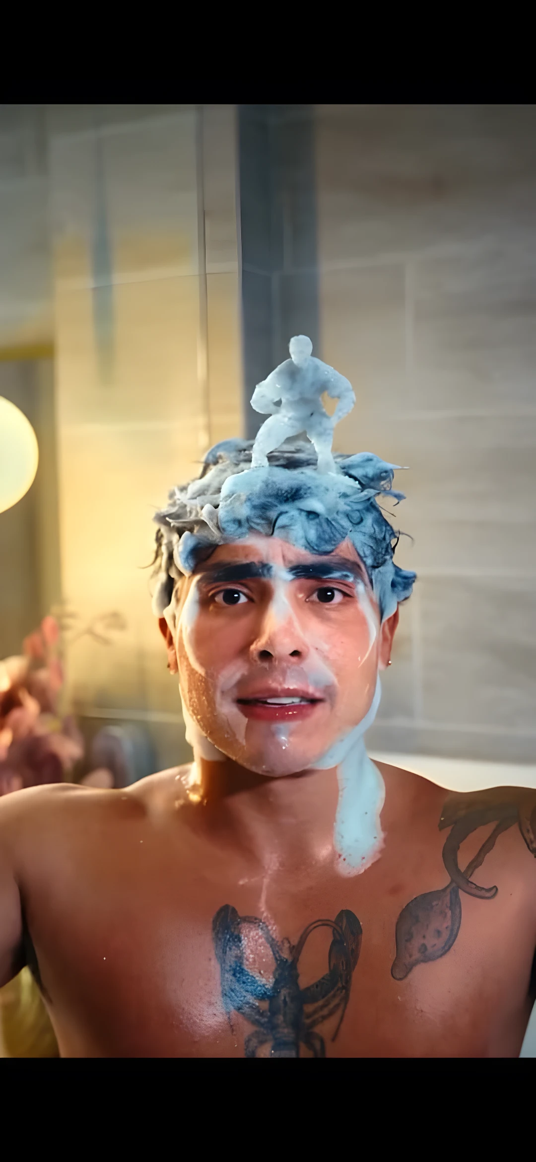 Hombre con espuma en la cabeza y un muñeco de jabón; tiene tatuajes de mariposa y ramas.