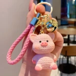 Llavero colgante de bolso escolar con forma de cerdito rosa texturizado, campanita dorada y correa trenzada.