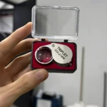 Lupa mini joyero plegable TRIPLE T 30X-21MM en estuche plástico transparente y forro rojo, sostenida en mano.