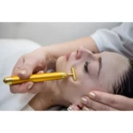 Masajeador facial T dorado en uso, aplicándose en la mejilla de una mujer, creando efecto tensor.