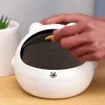 Mini basurera blanca con tapa estilo gato, manos depositando objeto en interior.